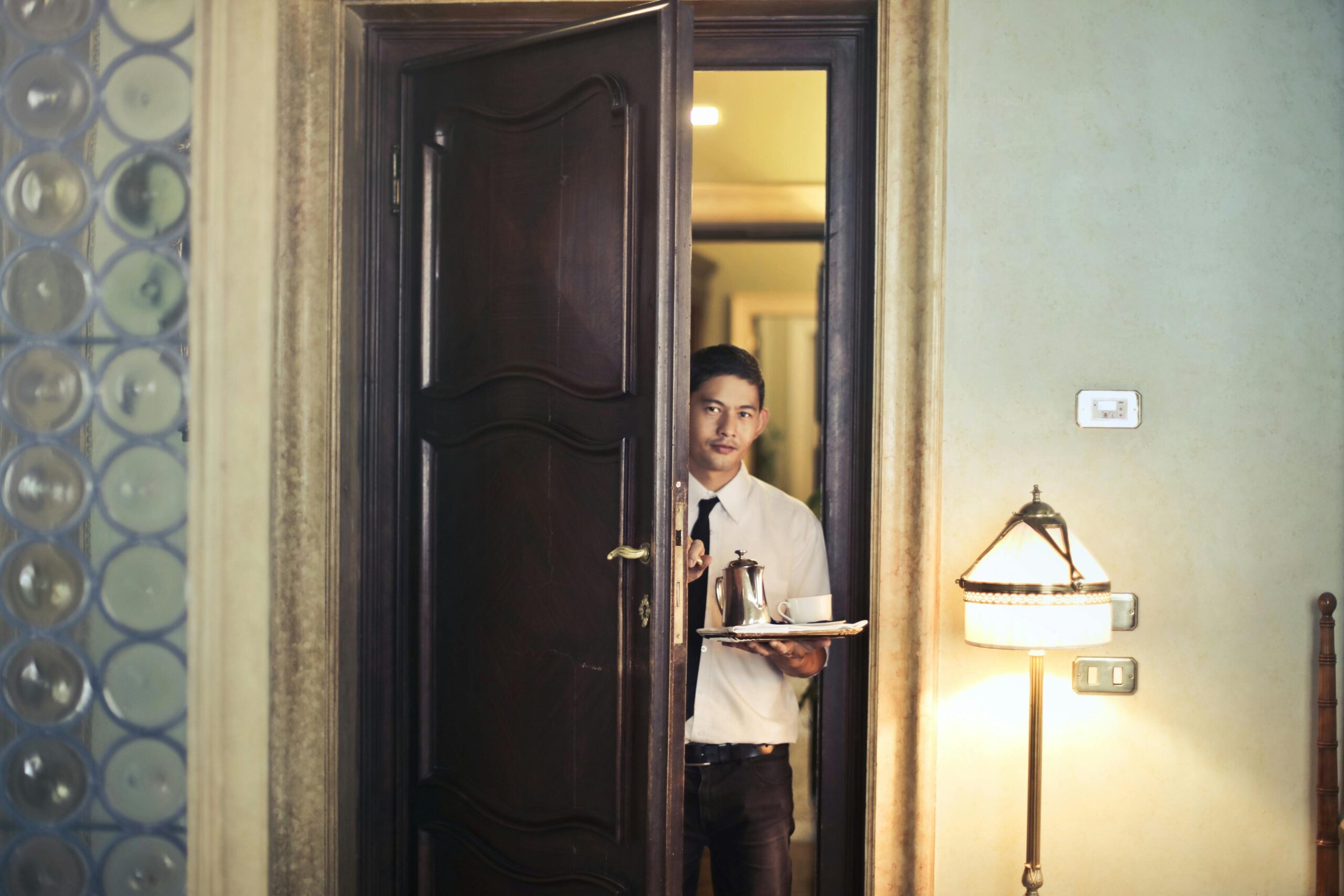 Young ethnic male room service waiter carrying tray with coffee pot while entering hotel room with stylish vintage interior